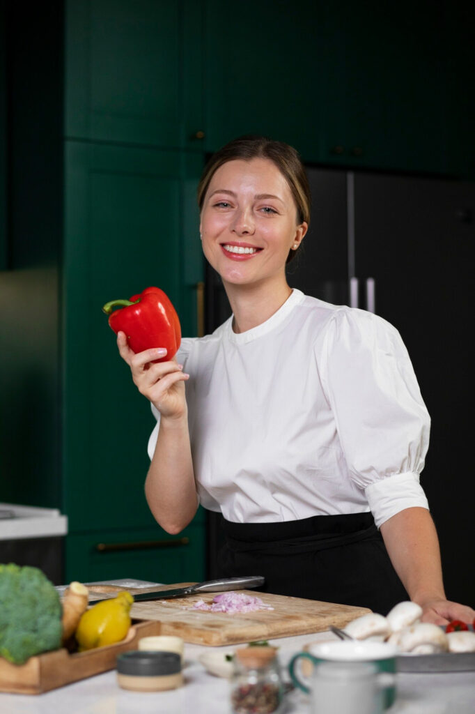 About me 1 Smiling woman chef Marina in a modern kitchen wearing a white chef coat, holding a wooden spoon and standing beside a bowl of fresh ingredients.